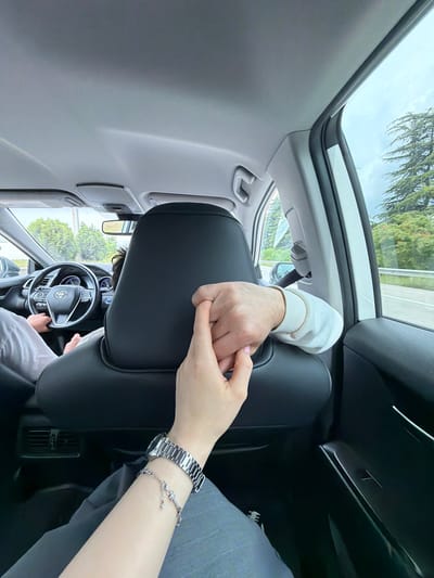 A view from inside a car, looking out through the windshield and sunroof, is dominated by the car's interior and the hands of two people holding each other. The driver can be seen holding the steering wheel, and the driver is also wearing a light-grey shirt. The hand of the passenger is holding the driver’s hand, and it is adorned with a silver watch and a silver charm bracelet. Outside, the landscape is composed of trees and a glimpse of the sky, indicating an outdoor setting. Given the specified city and country, it can be inferred that the car is within the vicinity of Tserovani, Georgia.