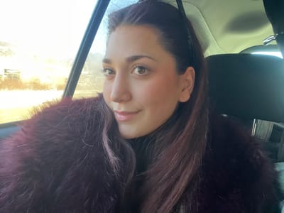 A female individual is depicted inside a vehicle, located in Dorlisheim, France. She occupies the left side of the frame, facing the camera with a slight smile. Her dark brown hair is styled in a ponytail, secured with a black hairband, and she has light brown eyes and a fair complexion. She wears a black turtleneck garment and a prominent dark plum-colored faux fur coat. The vehicle's interior features a light-colored ceiling and a black seat headrest visible to the subject's right. Through the left window, strong natural light indicative of low sun illuminates an indistinct outdoor environment, comprising blurred structures and potential vegetation, consistent with a moving vehicle during sunrise or sunset.