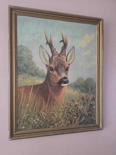 This is a framed painting of a deer's head and neck emerging from tall grass and wildflowers. The deer is brown with distinct antlers, one of which is partially obscured. The background features a soft blue sky with scattered clouds and a hint of a green landscape with trees. The overall scene appears to be outdoors, possibly a meadow or field, with a focus on the deer's alert gaze directed upwards and slightly to the left. The painting is housed in a simple gold-colored frame. There is no visible text in the image. The context provided indicates the location is Brzeg, Poland, but the painting itself does not offer specific cues for this. The lighting suggests it is daytime.