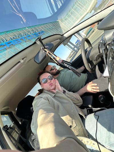 A selfie-style photo shows two people inside a vehicle. One person is seated behind the steering wheel and wearing a green shirt and sunglasses. The other person is in the passenger seat wearing a light-colored jacket and sunglasses and is turned towards the camera. Outside of the vehicle's windshield, a blue sign with Russian text and Cyrillic characters is visible. The name of the city, Ижевск (Izhevsk), Russia is written on the blue sign.