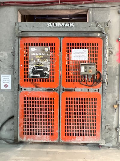This image displays an industrial construction hoist door, branded "ALIMAK," integrated into a concrete wall at a construction site in Aulnay-sous-Bois, France. The door comprises four rectangular, perforated orange metal panels within a grey metal frame. The upper left door panel features a safety poster titled "UN IMPRÉVU ? STOP!", depicting illustrated safety steps. The upper right panel bears a white sign in French, stating, "PLEASE REPORT TO THE SECURITY POST FOR THE OPENING OF THIS DOOR." Below this sign, a grey electrical control panel with green, black, and a red emergency stop button, along with associated black wiring, is mounted. A metal chain hangs from the right side of the door frame, and a yellow padlock secures a bracket on the adjacent concrete wall. To the left of the door, a circular sign with a crossed-out flame symbol prohibits "lift" use in case of fire. Faint grey markings resembling "H2O-S08" are visible on the concrete wall. Red electrical conduits are present above the hoist opening. The immediate ground in front of the door is a light grey concrete floor with minor debris. No individuals are present in the scene.