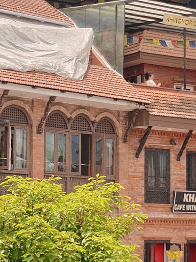 The image shows a close-up view of a red brick building with multiple windows and a terracotta tiled roof. The building has arched windows with decorative lattice work. In the foreground, there is lush green foliage from a tree. On the upper level of the building, a person is visible on a rooftop, and a sign that reads "KHASYO R CAFE WITH" is partially visible. Prayer flags are strung across the rooftops. A large white tarp covers a section of the roof. The overall scene appears to be an outdoor urban environment in Gokarneshwar, Nepal.