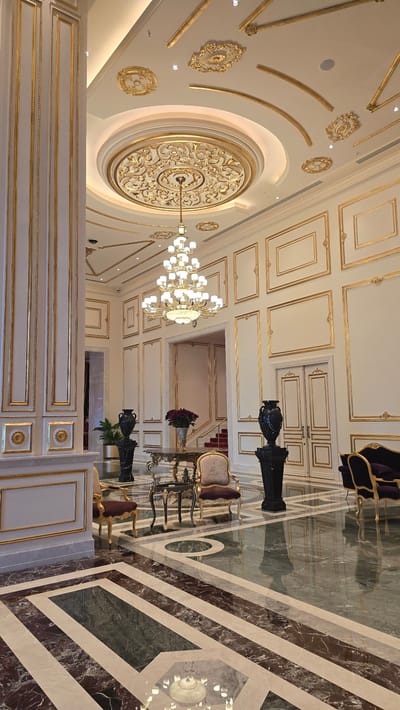 This image depicts the opulent interior of a lobby or reception area. The room is characterized by its lavish decoration, featuring extensive use of gold accents on white walls, columns, and the ceiling. A large, multi-tiered chandelier hangs from a circular, ornate ceiling medallion. The walls are paneled with gold trim framing rectangular sections, and decorative gold reliefs are affixed to the ceiling.

The flooring is a highly polished marble with a geometric pattern of contrasting colors, including dark brown, white, and green. A grand staircase with a red carpet is visible in the background, leading upwards.

Several pieces of furniture are placed in the room. Two ornate chairs with dark upholstery and gold frames are positioned around a decorative table. A dark, large vase stands on a pedestal to the right of the table, and another similar vase is visible further to the right. In the background, a dark purple sofa with gold trim is partially visible. Plants in dark pots are also present.

The lighting is warm and ambient, emanating from the chandelier and recessed ceiling lights. The overall impression is one of luxury and grandeur. There are no people visible in the image.

While the image showcases a specific interior design, the prompt requests that I provide details about the location in Agios Epiktitos, Northern Cyprus. However, the image itself contains no visual cues or information that would allow for the identification of its specific location within Agios Epiktitos or any other city in Cyprus. The description is limited to the visual elements present in the photograph.