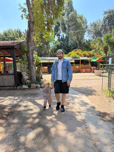 A man and a young child are walking together on a dirt path. The man is wearing a denim jacket, a white t-shirt, and black shorts. The child is wearing a brown shirt and pants. The background includes trees, a building, and fencing. The location is likely the city of Zvulun Regional Council, Israel.