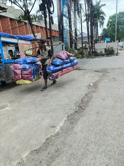 This image captures a street scene, likely in an urban or semi-urban area, during daylight hours. The focal point is an elderly man carrying a significant load of goods balanced on a shoulder pole.

The man, who appears to be a street vendor or peddler, is walking on a concrete or asphalt road. He is seen in profile, moving from left to right across the frame. He has dark skin, a mustache, and is wearing a dark cap, a light-colored short-sleeved shirt over a darker undershirt, dark trousers, and dark sneakers. A black fanny pack or waist bag is visible around his waist. He uses both hands to steady the pole and the suspended loads.

The goods he carries are brightly colored, wrapped in plastic, and appear to be soft items like pillows, cushions, or rolled mats. On his left side, there are several rectangular and square-shaped items, predominantly blue, pink/red, and yellow, stacked and tied. On his right side, similar blue and pink/red items are carried, including at least two long, rolled items that are pink/red and blue, suggesting they might be floor mats or sleeping mats. These items are suspended from the shoulder pole by ropes.

To the left of the man, partially visible, is a street food cart. It has a blue canopy and a metal frame. Inside the cart, a young person or child with dark hair is visible, looking towards the right. The cart appears to have food containers or dishes. Some text is visible on a banner or sign attached to the cart, with "Mie Ayam" clearly readable, which suggests it sells chicken noodles, a common street food. Below "Mie Ayam," the word "Mari" is also visible. Another bag of yellowish items is seen on the counter of the cart.

The background consists of several elements. Behind the man and the cart, there is a building with an orange-brown brick-like facade and large windows or openings. Numerous utility poles with a dense network of black cables crisscross the mid-ground. Blue scaffolding or netting is visible around some of the structures in the background, suggesting ongoing construction or renovation. Further back, palm trees and other green foliage are visible, indicating an outdoor setting with some greenery. The sky appears overcast or a bright, diffused light.

The road surface is made of concrete or asphalt and shows signs of wear and tear, with cracks and uneven patches, particularly on the right side of the image where a prominent crack runs diagonally. Some small debris is scattered on the ground. The overall impression is one of daily life and labor in a bustling, somewhat weathered urban environment.