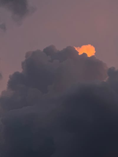 This is a close-up, vertical photograph of dark, moody clouds against a dusky purple sky. A small patch of cloud near the top center is brilliantly illuminated by a warm, orange glow, suggesting the sun is setting or has just risen behind it. The rest of the clouds are rendered in deep grays and purples, creating a dramatic and atmospheric effect. The image is devoid of any human elements, structures, or any indications of a specific location within Savenki, Russia. The focus is solely on the celestial phenomenon.