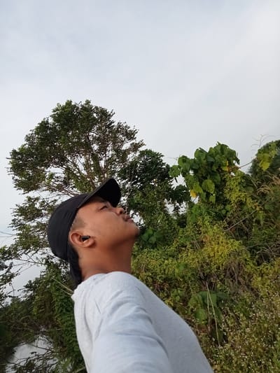 An outdoor, low-angle, eye-level medium shot shows a young man wearing a black baseball cap and a light grey t-shirt. He is looking up and to the right, with a black earbud in his right ear. A large green tree is behind him and to his left, with a dense thicket of green foliage and some small white flowers filling the right side of the frame. The sky is overcast and a pale grey. The environment suggests a rural or natural setting within Teluk Kenidai, Indonesia.