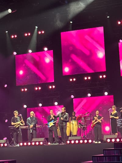 This media file captures a live musical performance taking place on a stage. The subjects are a band of musicians. The setting is a concert hall or large indoor venue, characterized by dark walls and a stage adorned with large, bright pink screens displaying abstract patterns and lighting effects. The event is a musical performance, with band members actively playing instruments.

The band consists of several individuals, including guitarists, a bassist, and a percussionist playing congas. They are dressed in casual, dark attire. One individual is singing into a microphone, while others are focused on their instruments. A woman in a black dress is also present, standing and possibly singing or addressing the audience. The stage is illuminated by spotlights and the large pink screens, creating a vibrant and energetic atmosphere. The time of day is evening or night, as indicated by the dark surroundings and stage lighting. There are no overt indications of weather.

Notable details include the presence of multiple microphones, musical instruments such as guitars, a bass guitar, and conga drums, and a drum kit visible in the background. The lighting is dynamic, with spotlights casting beams of light and the pink screens providing a colorful backdrop. Some of the lighting appears to be in a spotlight beam creating smoke-like effects. The emotions conveyed are those of performance energy and engagement with the music.

Visible text is present on a banner above the stage, but it is partially obscured and difficult to read clearly, though it appears to be in Cyrillic script. The location is identified as Nemchinovka, Russia.