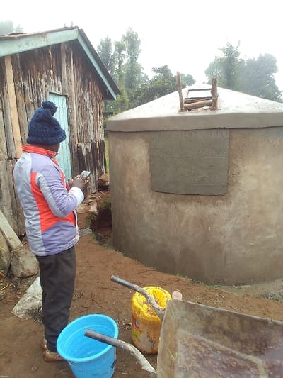 The image depicts an outdoor scene in Ihururu, Kenya, on a damp, overcast day, characterized by reddish-brown earth and dense green foliage in the background.

A lone individual, viewed from the rear, stands slightly left of center, dressed in a dark blue knitted beanie, a multi-paneled jacket (grey, white, orange, and purple sections), dark trousers, and light-colored open-toed footwear. The person is actively engaged with a smartphone, holding it with both hands at chest level.

To the immediate left of the individual is a rustic, single-story wooden building constructed from vertical planks, featuring a gabled roof with a light blue or green fascia and a closed wooden door on its visible side.

Dominating the right side of the frame is a large, grey, cylindrical concrete water tank with a conical top. An apparent access hatch on the tank's apex is fitted with a simple wooden ladder-like structure. A rectangular inset plaque on the tank's side contains embossed text, partially legible as "JESPELLA PROJUCT", "IHURURU KENYA", "AFRICAN", and "CEMENT TANK".

In the foreground, a blue plastic bucket, a yellow plastic container, and the rusty metal basin of a wheelbarrow are visible on the ground. The ground between the person and the tank shows signs of slight excavation or a natural depression.