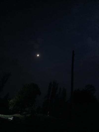 The image depicts a nighttime scene with a crescent moon visible in the dark sky. Silhouetted trees and a utility pole are present in the foreground. On the ground, a white object, possibly a vehicle or structure, is partially visible. The overall environment appears to be rural or semi-rural, with no specific landmarks indicating it is within the city of Auk, Kyrgyzstan.