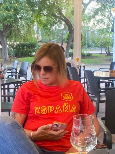 A woman is seated at an outdoor table at what appears to be a cafe or restaurant in San Pedro De Alcántara, Spain. She is wearing a red t-shirt with "ESPAÑA" and a crest printed in yellow on the front, and blue jeans. She has sunglasses on and is holding a pink smartphone in her hands. In front of her is an empty stemmed beer glass with remnants of foam. The background shows trees, other tables and chairs, and buildings.