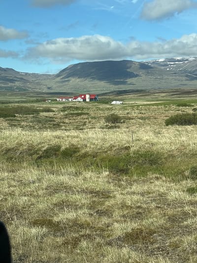 A panoramic view of a rural landscape in Breiðabolsstaðir, Iceland, is depicted under a partially cloudy blue sky. In the mid-ground, a cluster of white farm buildings with red roofs is situated in a green and brown field. A long, white trailer or vehicle is parked near the buildings. The foreground is dominated by dry, tall, golden-brown grass, interspersed with patches of darker green vegetation and a thin wooden fence post. In the background, rolling hills and mountains rise, some with patches of snow on their peaks. The overall scene suggests a remote agricultural setting in a mountainous region.
