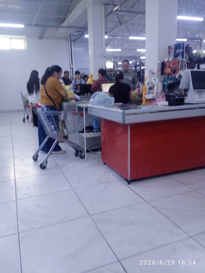 The image shows the interior of a brightly lit supermarket in Ak-Suu, Kyrgyzstan. In the foreground, a red checkout counter with a silver trim is visible. A female cashier, wearing a black shirt, is operating the register. Behind her, a man in a grey t-shirt, presumably a customer, is standing. To the left of the counter, a woman in an orange shirt and blue pants is pushing a shopping cart. Another woman with long dark hair is standing behind her. Several other shoppers and employees are visible in the background, some interacting with each other. The floor is tiled with large light-colored tiles. The timestamp "2026/6/29 16:34" is present in the bottom right corner of the image.