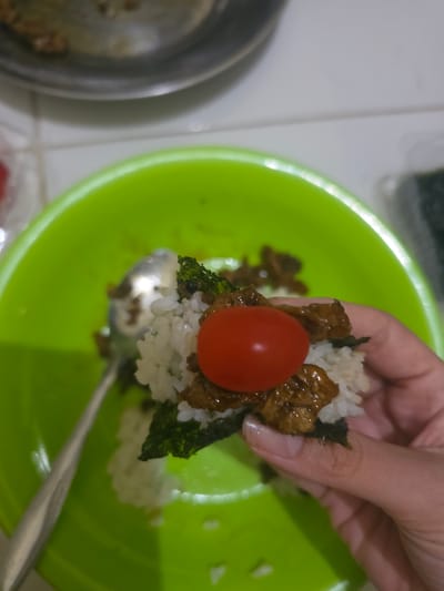 A hand is holding a small rice ball or onigiri topped with a red cherry tomato. The rice ball also contains nori seaweed and some cooked meat. The onigiri is being held over a bright green bowl, which contains more rice and bits of meat and seaweed. A spoon is also in the bowl. In the background, there's a metal bowl containing more food. The image appears to be taken in a domestic kitchen environment. The location, Pintomakmur, Indonesia, is stated in the instructions but not evident from the image content.