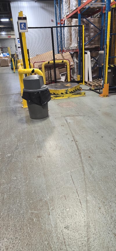 This image depicts an indoor industrial or warehouse setting, likely in Drummondville, Canada, characterized by concrete flooring, tall shelving units laden with boxed goods, and yellow safety barriers. In the foreground, a grey, conical trash receptacle with a black liner is visible. To its right, a yellow-and-black striped platform, possibly a scale or a section of a conveyor system, is present, partially enclosed by a black mesh fence. A yellow metal stanchion with a sign displaying a wheelchair accessibility symbol and a figure pushing a broom stands to the left of the trash can. The environment appears to be a functional workspace, with no individuals directly observed within the frame, but the presence of safety equipment and industrial elements suggests ongoing operations.