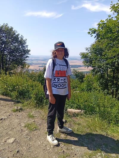 A young person stands on a dirt path, facing the camera, with a scenic view of fields and trees in the background. The person is wearing a black baseball cap, a white t-shirt with a graphic print, black pants, and white sneakers. A black backpack is visible over their shoulders. The path is rocky and dusty, with green grass and shrubs growing alongside it. In the background, rolling hills and farmland stretch towards the horizon under a clear blue sky with scattered clouds. The location is identified as being in the city of Sobótka, Poland.