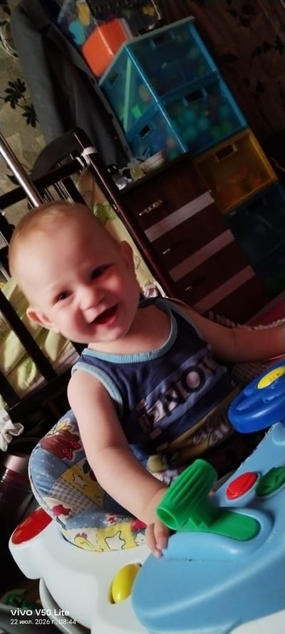 A baby is sitting in a walker with a toy console in front of it. The baby has blonde hair and is smiling. The walker is blue and white with a colorful seat. The background is blurred, but it appears to be a room with toys and furniture. A watermark on the bottom left corner indicates the photo was taken with a "vivo V50 Lite" phone on "July 22, 2026, 08:44" in "Imeni Sverdlova, Russia."