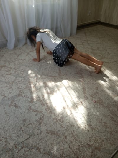 A young girl is performing a plank exercise on a patterned carpet. She is wearing a white t-shirt and a dark skirt with white stars. Sunlight streams through sheer curtains, creating a dappled light pattern on the floor. The room appears to be a bedroom or living area. The location is stated to be Кенжекөл, Kazakhstan.