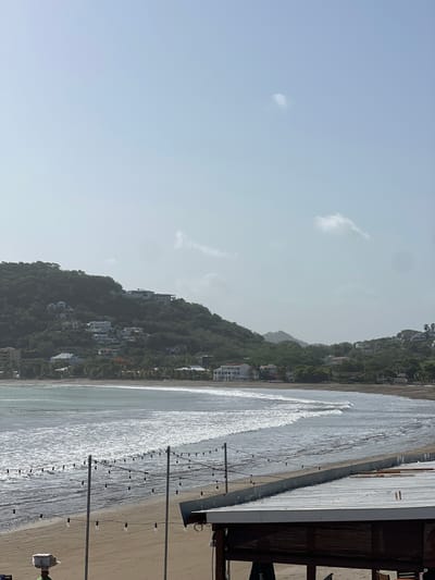 The image depicts a beach scene in San Juan del Sur, Nicaragua. In the foreground, a covered structure with a corrugated metal roof is visible, along with strings of cafe lights extending across the beach. The sand is empty except for a person walking with a package on their head. The ocean is calm with gentle waves breaking on the shore. In the background, a lush green hillside is dotted with white houses, some of which are large villas. The sky is hazy and light blue with a few wispy clouds. The overall impression is a tranquil, tropical beach setting.