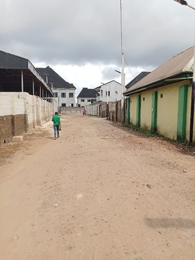 The image shows a dirt road in Ovwian, Nigeria, located at coordinates 5.485 latitude, 5.806 longitude. The road is lined on both sides by buildings, with a white concrete block wall on the left and a light yellow building with green doors on the right. The road is empty, except for one man walking toward the camera in the center of the frame. There is a second person walking in the distance to the left. The sky is overcast, and the overall lighting is hazy. The buildings appear to be unfinished or in the process of being renovated, with some missing siding. There is a power line running down the center of the road and some weeds growing along the sides. The scene is somewhat desolate and gives the impression of a rural area still under development.