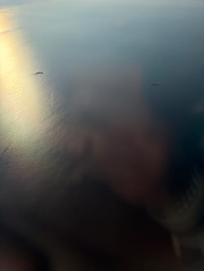 The aerial photograph displays a vast expanse of ocean, with two cargo ships visible in the distance. The sun glints off the water in the upper left quadrant, casting a bright, shimmering light. The lower portion of the image is obscured by a blur, possibly from the camera lens or an obstruction. The overall scene is dominated by muted blues and grays, with the bright sunlight offering a stark contrast. The context of Çiftlikköy, Turkey, suggests this is likely a view over the Sea of Marmara or the coastline near the town, known for its ports and maritime activity.