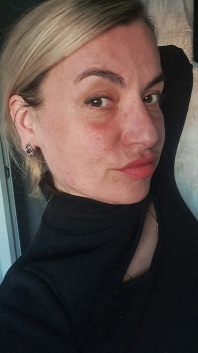 A woman is pictured in a close-up shot, facing slightly to the right. She has fair skin, brown eyes, and blonde hair. Her lips are painted with red lipstick. She is wearing a black turtleneck sweater and an earring on her left ear. Part of a chain necklace is visible at her neck. The background appears to be a wall or a surface with a light-colored pattern. There is no indication of a specific location in Меткомелино, Russia.