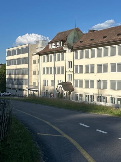 The image depicts a building with a brown tiled roof and a green-domed structure, likely an older section of the building. The building facade is light yellow with numerous windows, many featuring blinds. One visible sign reads "BLANCPAIN". The building appears to be a factory or office complex, possibly from the early to mid-20th century, as indicated by the number "1930" engraved on the facade. Adjacent to the building is a paved road with yellow and white markings. Tall grass and vegetation line the roadside and the building. A clear blue sky with a large white cloud is visible. The sun appears to be shining, suggesting daytime. The scene is static, with no people or overt activity captured, except for a white vehicle with some text visible on its side parked on the side of the road in the distance. The overall setting is industrial or commercial, set against a natural backdrop. The crops highlight different sections of the building, the road, and the sky. The location context identifies the scene as Le Sentier, Switzerland.