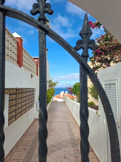 The image shows a narrow pathway in a residential area, likely in Santiago del Teide, Spain, viewed through a black wrought-iron gate. The pathway is paved with terracotta tiles and flanked by white walls. On the left wall, a row of mailboxes is visible. The pathway leads towards the ocean, which is visible in the distance under a blue sky with scattered clouds. Greenery and pink bougainvillea flowers adorn the right side of the path. The overall impression is of a sunny, coastal environment.