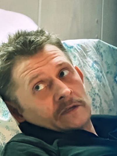 The image shows a close-up, angled shot of a man lying down, looking to his left. He has short, spiky brown hair and a mustache. His eyes are wide and his eyebrows are furrowed. He appears to be wearing a dark-colored shirt or jacket. The background consists of a pillow with a floral pattern and a plain, light-colored wall. There is no visible indication of the city of Dondușeni, Moldova, or any specific urban environment. The scene is sterile and lacks any contextual clues about its location.