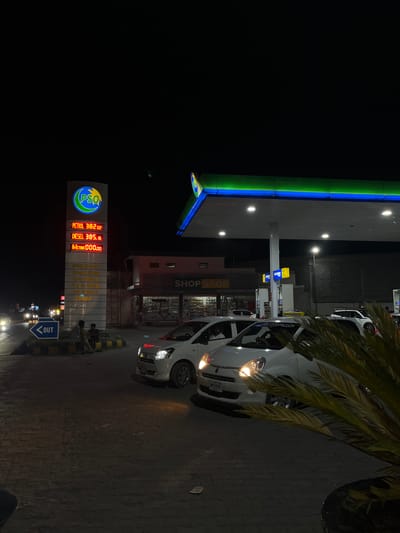 This is a nighttime photograph of a PSO (Pakistan State Oil) gas station in Kabal, Pakistan. The scene is dominated by a brightly lit vertical price board displaying "PETROL 382.02," "DIESEL 385.16," and "HI-OCTANE 000.00." Below the fuel prices, a list of services includes "PSO Cards," "Shop Stop," "Car Wash," "Tyre Shop," "24 Hours," and "Masjid."
To the right of the price board, a gas station canopy with blue and green illuminated trim covers several fuel pumps. Under the canopy, multiple white cars are parked or queuing for fuel. The closest white car on the right has its headlights on, and a person is visible in the driver's seat. Its license plate, "SWT B 0748," is partially visible. Further to the right, a potted plant with green palm-like foliage partially obscures the vehicle.
Behind the price board and pumps, a single-story building labeled "SHOP STOP" serves as a convenience store. Another building with a corrugated block wall is visible behind the pumps on the far right.
On the left side of the frame, near the base of the price board, three men are seated on a low concrete barrier or curb. A blue directional sign with "OUT" in white text points left, indicating an exit from the station. The ground is paved with dark interlocking bricks. Distant vehicle headlights are visible further down the road on the left, indicating other traffic. The overall illumination comes from the station's overhead lights and signage against a dark sky.