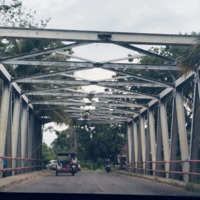 The image provides a first-person perspective from within a vehicle traveling through a metal truss bridge in Teupinkupula, Indonesia, on an overcast day. The scene is dominated by the bridge's intricate grey metal framework, composed of a series of intersecting diagonal and horizontal beams that form a tunnel-like structure over the road.

The paved road is visible leading through the bridge, flanked by red safety railings on both sides. Beyond the bridge's metal lattice, abundant green tropical foliage, including large trees and palm fronds, suggests a lush, possibly rural or semi-rural, environment. The sky is a uniform, light grey, indicating an overcast weather condition.

Vehicles are present on the road ahead. Directly in front, slightly to the left of the center, is a dark-colored three-wheeled vehicle, possibly an auto-rickshaw or a customized scooter with a passenger cab. Further down the road on the right, a motorcycle is visible. A white car is partially discernible behind the three-wheeled vehicle, to the left. The activity appears to be routine travel across the bridge. No specific emotions are conveyed, and no visible text is discernible on the bridge or vehicles.
