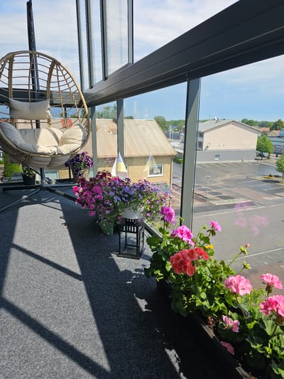 The image depicts a covered balcony or sunroom in a residential building in Smålandsstenar, Sweden. A light-colored rattan hanging chair with cushions is positioned on the left side of the frame. In front of the chair, a row of potted plants with purple and pink flowers is arranged along the balcony railing. To the right, a planter box filled with pink and red geraniums is visible. The balcony has a grey carpeted floor, and sunlight casts geometric shadows across it. Through the glass windows, an outdoor scene is visible, including buildings, a parking lot, and trees. The buildings appear to be of various sizes and architectural styles, some with light-colored facades and corrugated roofs. The sky is blue with some scattered clouds. The overall impression is one of a well-maintained private outdoor space overlooking a typical urban or suburban environment.