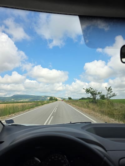 The image is a first-person view from inside a vehicle, looking forward through the windshield. The vehicle is on a paved road with white dashed lines marking the lanes. The road stretches into the distance and is flanked by dry grass and fields on either side. In the background, there are rolling green hills under a bright blue sky filled with fluffy white clouds. A single red car is visible in the distance on the road. The interior of the vehicle is visible in the foreground, including the top of the dashboard and a portion of the steering wheel. The scene appears to be in a rural area. The image is not located in Medovnitsa, Bulgaria.