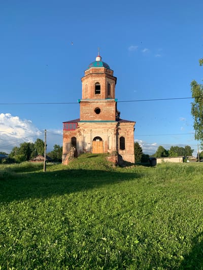 This is a full-frame, eye-level, outdoor photograph of an old, red brick church in the village of Svyatitsa, Russia, on a clear, sunny day. The church is the central subject, with its octagonal bell tower topped by a teal-colored dome with a cross. The church has arched windows and a wooden door. The building appears aged, with some signs of disrepair, particularly at the base where some bricks are missing or crumbling. In the foreground, there is a large expanse of green grass, with a shadow cast by an unseen object to the left. In the background, there are trees, some wooden buildings, and a power line stretching across the frame. The sky is a vibrant blue with a few scattered white clouds and numerous small, dark birds flying.