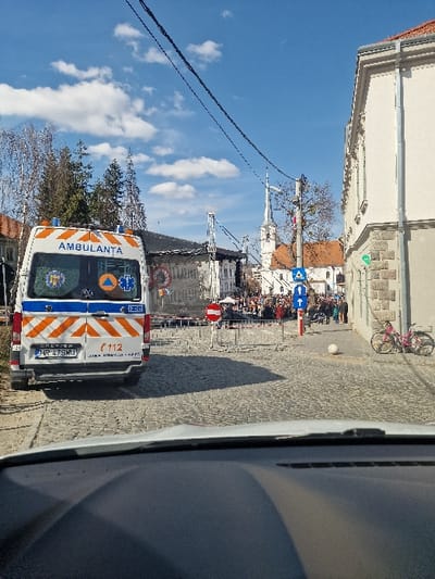 The image depicts a daytime street scene in Odorheiu Secuiesc, Romania, captured from within a vehicle, with a clear blue sky and scattered white clouds overhead. The perspective is directed towards what appears to be Piața Marton Áron, with the Reformed Church steeple visible in the background.
In the immediate foreground, the upper portion of a vehicle's dashboard is visible. The street, composed of cobblestones, extends into the midground. A white ambulance with orange reflective markings, displaying "AMBULANTA" and the emergency number "112," is parked on the left, facing away from the viewer. Its license plate reads HR 47 SMU.
Further down the street, temporary barriers and a red circular "no entry" sign restrict access to a gathered crowd of people. Behind the crowd, a large metallic scaffolding structure, indicative of a stage or temporary installation, is prominent. To the right side of the street, a light-colored multi-story building with a stone base is visible, next to which a pink bicycle is parked. A white spherical object rests on the cobblestone surface near this building.
In the background, a distinct white church steeple with a pointed spire rises above other buildings, one of which features a terracotta-colored roof. Overhead, multiple black power lines traverse the frame horizontally. Bare deciduous trees and evergreen trees are visible on the left side of the scene.
