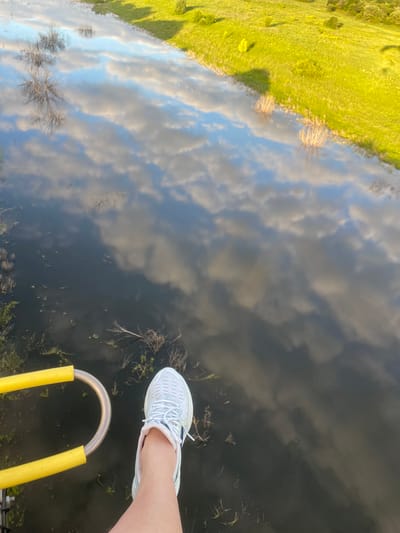 The image shows a first-person perspective of a person's leg and foot wearing a white sneaker, positioned next to a yellow handrail. The scene appears to be over a body of dark water, reflecting the sky with white clouds and a green grassy embankment with trees. This reflection suggests the vantage point is elevated, possibly from a hot air balloon or a bridge. The location is identified as Турово, Russia. The environment is natural, with water and vegetation. There are no other people clearly visible.