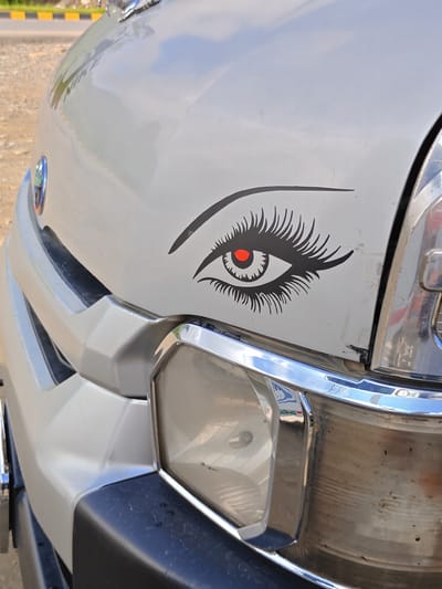 A close-up, eye-level shot of the front of a white vehicle shows a black sticker of a stylized eye with long eyelashes and a red pupil on the hood. To the right of the sticker, a portion of a chrome grille and a headlight assembly is visible. The vehicle is parked outdoors, with a dirt shoulder and a paved road with yellow and black markings in the background, suggesting a location on a street or roadside in Tharpu, Nepal. The lighting indicates it is daytime, and the overall condition of the vehicle's front suggests it has been used.