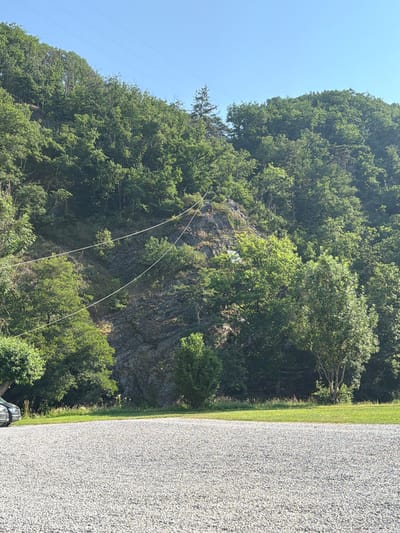 The image depicts a rocky, tree-covered hillside under a clear blue sky, with a gravel path running along the right side. The scene appears to be set in a rural or natural environment, possibly a park or hiking area, given the lush greenery and rocky terrain. A few thin, white lines, possibly wires or cables, stretch across the hillside, disappearing into the trees. The lighting suggests it's daytime, likely a sunny day. The overall mood is serene and natural. There is no visible text in the image.