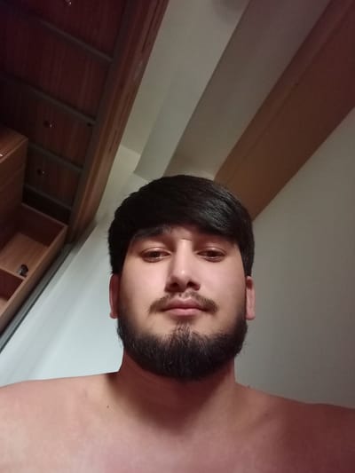 A selfie is captured from a low angle, showing a young man with dark hair and a beard looking directly at the camera. He is shirtless, and his shoulders and upper chest are visible. Behind him, a wooden wardrobe with open shelves is partially visible on the left. The background is a light-colored wall with vertical wooden beams. The image does not contain information about the city of Hongshan District, China.