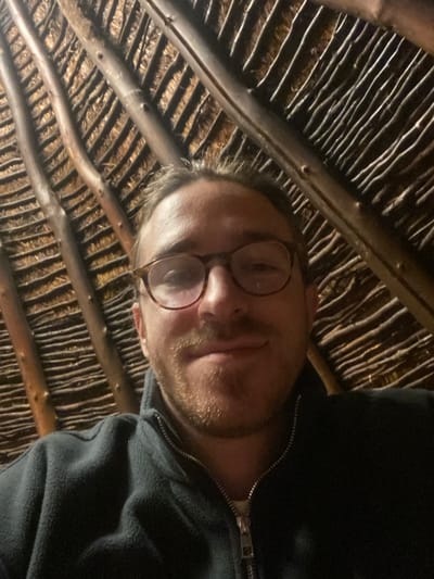 A male individual is depicted from the upper chest upwards in a close-up, centered perspective. He has light brown hair, a short beard, and wears tortoiseshell-rimmed glasses, exhibiting a slight closed-lip smile while looking directly into the camera. He is dressed in a dark blue fleece jacket with a visible zipper pull. The background consists of an interior ceiling constructed from a network of dark, substantial wooden beams arranged diagonally, overlaid with numerous lighter, thinner horizontal wooden rods or reeds, creating a distinct rustic, woven pattern. The illumination is low, originating from an unseen source. This scene is situated in Dulovine, Montenegro.