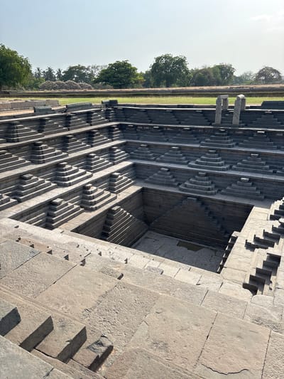 The image shows the stepped tank of Pushkarani in Kamalapura, India. The tank is made of stone and features a series of stepped levels leading down to a square-shaped center. Along the side of the tank is a grass strip, a low stone wall, and a line of trees. The sky is pale blue. The tank's architecture features repetitive geometric patterns, creating a complex interplay of light and shadow. The stone used in the structure appears aged and weathered.