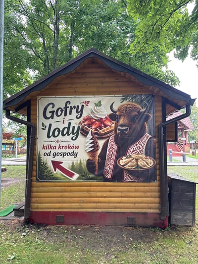 A wooden kiosk with a prominent banner advertising "Gofry i lody" (waffles and ice cream) stands in an outdoor setting, likely within the Bialowieza National Park in Bialowieza, Poland. The banner features a stylized illustration of a European bison wearing traditional clothing and holding a plate of pierogi. Accompanying the bison are images of a waffle topped with strawberries and whipped cream, and a swirl of ice cream. Text below the images states "kilka kroków od gospody," meaning "a few steps from the inn." The kiosk is surrounded by greenery, with trees in the background and grass in the foreground. Other structures, including a covered seating area and a small wooden shed, are visible in the distance. The overall scene suggests a tourist-oriented establishment offering local food items.