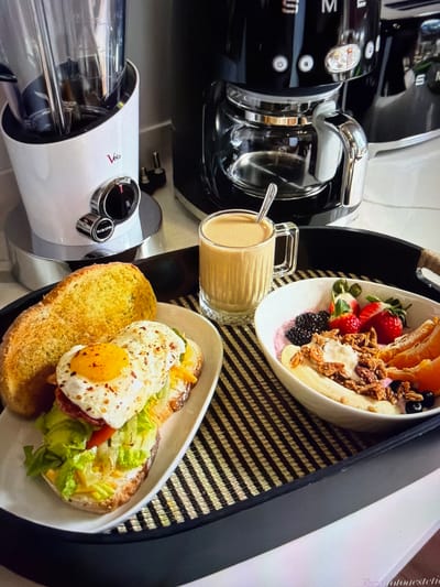 This image presents a vibrant and appetizing breakfast spread arranged on a black serving tray, which features a distinctive black and brown striped woven mat. The meal includes a bowl of fresh fruit, yogurt, and granola, with visible strawberries, blackberries, orange slices, and banana chunks artfully placed. Alongside, a clear glass mug holds a light brown beverage, possibly coffee with milk or a latte, with a spoon inside. A savory component consists of a piece of toasted bread topped with a perfectly cooked sunny-side-up egg, lightly sprinkled with spices, accompanied by fresh green lettuce and other elements like red pepper.

In the background and to the left, several modern kitchen appliances are visible. A glossy black Smeg espresso machine with chrome accents is prominent, displaying "SMEG" branding and button controls, including a power symbol. Below it, a white appliance, possibly a blender or juicer, features a clear processing chamber, chrome dials, and the stylized red logo "Vela" (or Velo) along with an "ON/OFF" indication.

The scene suggests a leisurely and well-prepared breakfast or brunch, creating a feeling of comfort and indulgence. The precise time of day is morning or early afternoon, consistent with breakfast items. Faint text, potentially a watermark or photographer credit like "Real Fotoestetic," is discernible on the far right edge. The provided context states the location is Ros Na Rí, Ireland.