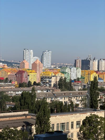 The image shows a panoramic view of the city of Kyiv, Ukraine, on a clear, sunny day. In the foreground, there are several multi-story residential buildings, some with flat roofs and others with pitched roofs. These buildings appear to be older Soviet-era apartment blocks. Further in the midground and background, there is a striking contrast with a cluster of brightly colored, modern residential buildings with pitched roofs painted in vibrant shades of pink, yellow, green, and blue. Behind these colorful buildings, taller, contemporary high-rise apartment buildings dominate the skyline, some with glass facades. In the far distance, a faint silhouette of other city structures and hills can be seen. The sky is a clear, pale blue with a few distant, small birds. There are no people or vehicles visible in the frame. The overall impression is one of urban development, with older and newer architectural styles coexisting. The scene appears to be a residential area within Kyiv, likely showcasing the "Comfort Town" residential complex, known for its distinctively colored buildings.