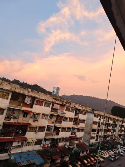 This image, captured in the Ampang Jaya Municipal Council, Malaysia, offers a view of a low-cost apartment complex bathed in the warm hues of a sunset. The sky is a gradient of pale blue and soft pink, dotted with scattered clouds. In the foreground, the multi-story apartment building exhibits signs of age, with weathered concrete facades and numerous windows, many with metal grilles. Various awnings and small balconies jut out from the structure. Laundry hangs from some of the balconies, and satellite dishes are visible on the exterior. The building is oriented at an angle, suggesting a tilted camera perspective. To the left of the building, a dense line of trees and a hillside form the background, with a modern high-rise building peeking out from behind them on the right side of the hill. Below the apartment complex, a parking lot is filled with a variety of cars. A tall, thin utility pole or antenna stretches vertically through the frame, partially obscuring the view of the sky. The overall scene suggests a residential area in Ampang Jaya during twilight.