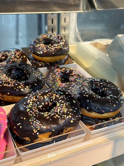 A close-up, eye-level shot shows an assortment of chocolate-frosted donuts topped with colorful sprinkles arranged in white paper liners, inside a display case. The donuts are golden brown on the bottom with a thick layer of dark chocolate frosting. The sprinkles are varied in color, including pink, blue, yellow, and green. In the foreground, a portion of a pink frosted donut is visible on the far left. In the background, blurred elements of a store interior are visible, including shelving and what appears to be other food items. The lighting is bright, casting some reflections on the donuts and the display case. There is no indication of Phu Thuy, Vietnam in the image.