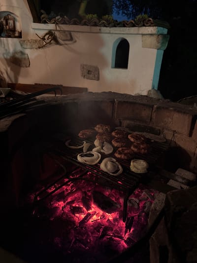 The image depicts an outdoor barbecue scene at night, set against the backdrop of a white-washed building with arched windows and terracotta roof tiles, characteristic of traditional architecture found in Taban, Bulgaria. The foreground is dominated by a brick barbecue pit filled with glowing red embers, illuminating a grill laden with sizzling burgers and thick-cut onion rings. Several pairs of tongs are visible on the rim of the pit, suggesting active food preparation. The surrounding environment is dark, with hints of foliage in the background, indicating a rural or semi-rural setting. The overall atmosphere is one of rustic culinary activity under the night sky.