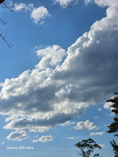 The image displays a bright blue sky filled with a mix of fluffy white and dark gray clouds. The clouds are dramatically arranged, with a large band of darker clouds dominating the upper right portion of the frame, casting shadows on the lighter clouds below. In the bottom right corner, the dark green foliage of a tree is visible, along with a portion of another tree in the upper right. The lower part of the image shows a thin black wire stretching horizontally across, with a small dark bird perched on it towards the right side. In the bottom left corner, the text "Galaxy S26 Ultra" is superimposed on the image. The scene is set outdoors, with the sky and clouds suggesting a dynamic weather pattern, possibly on the outskirts of West Hartford, Connecticut.