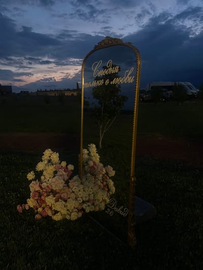 In the city of Sasunik, Armenia, a reflective mirror with an ornate gold frame stands outdoors at dusk. The mirror is adorned with white and pink flowers at its base, and text is inscribed on its surface in a script font. The reflected text reads "Сегодня только о любви," which translates to "Today is only about love." Below this, the name "Давид" and the number "26" are also visible on the mirror. The background shows a cloudy sky with hues of orange and purple, suggesting sunset. In the distance, there are trees, some buildings, and vehicles, including vans and cars. The mirror is positioned on a grassy area, with a dirt path running behind it. The overall atmosphere is serene and contemplative.