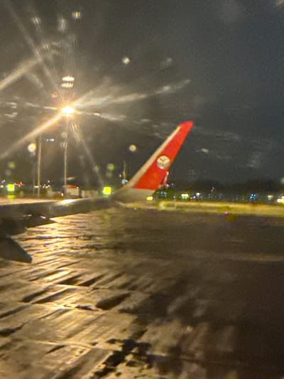 The image depicts a nighttime scene from inside an aircraft, looking out onto a wet airport tarmac. In the lower-left foreground, a portion of an airplane wing is visible. To the right, the prominent red vertical stabilizer (tail fin) of another aircraft is seen, featuring a white triangular section near the top with a circular logo, identifiable as the stylized phoenix of Air China. The tarmac below is highly reflective, indicating wet conditions, likely from rain, with light sources creating streaks and reflections across its textured surface.

Multiple bright artificial lights illuminate the scene. A prominent, starburst-effect light source is centrally located in the upper-left, casting strong illumination and lens flares. In the mid-ground and background, numerous smaller, colored airport lights (yellow, green, and white) are visible, outlining taxiways or runways and other airport infrastructure. The sky above is dark, confirming the nighttime setting. There are no people visible in the frame.

Given the Air China livery and the request for location, this scene is consistent with an airport tarmac in Jianyang City, China, specifically Chengdu Tianfu International Airport (TFU).